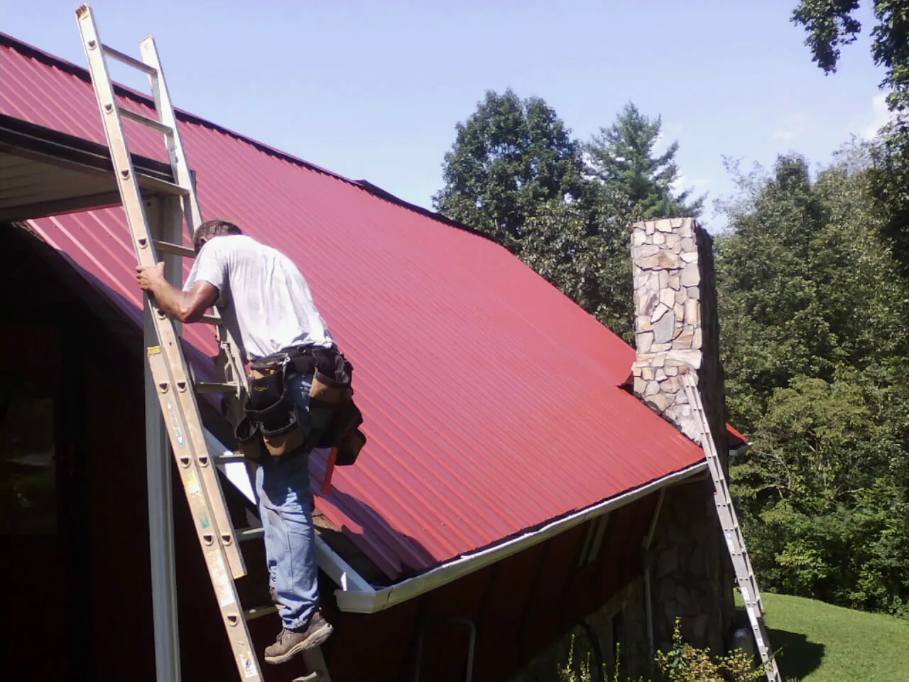 Roofer on a ladder beside a red metal roof and stone chimney — Soffit & Fascia Repair by Roofing Brothers in Twinsburg